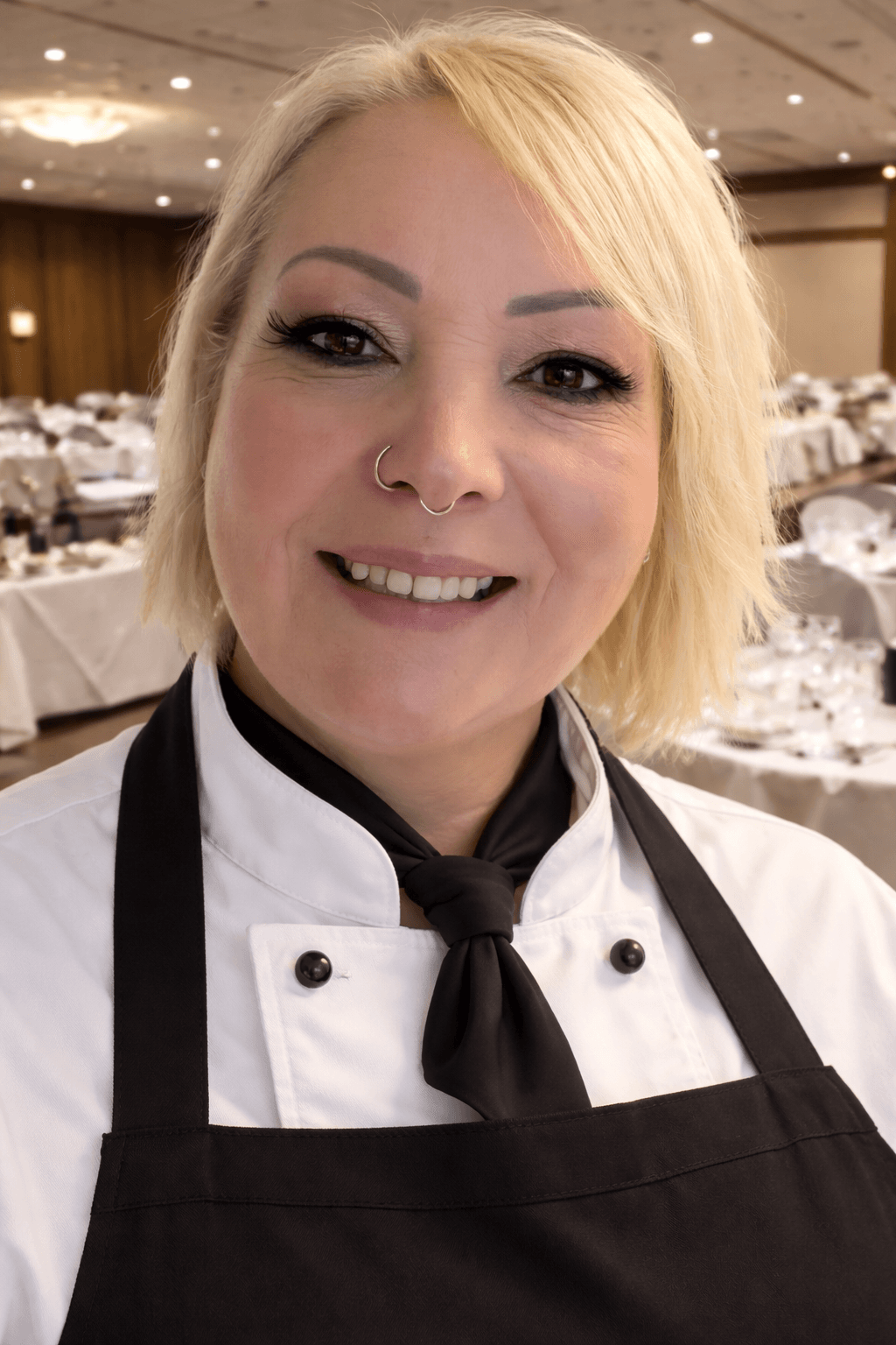 Smiling blonde woman in a white chef coat and black apron in a banquet hall.